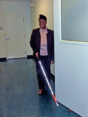 Two photos show a woman walking along a hall, approaching the edge of an open door.  Her left hand trails the wall and her right hand holds a cane with the tip on the floor straight ahead of her hand.  The cane never touches the door, and she is about to collide into it.